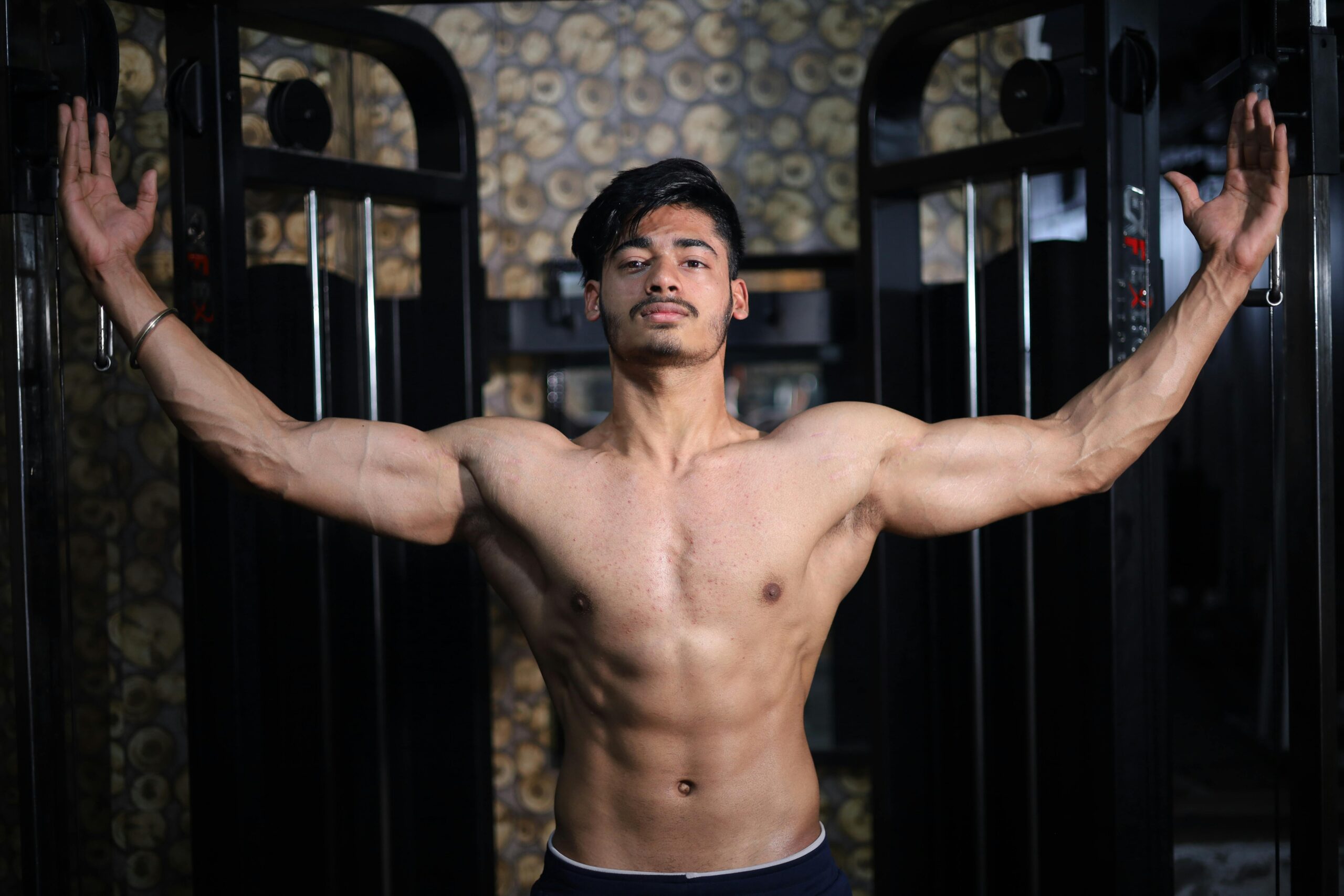 A shirtless South Asian man shows off his muscular physique while posing in a gym setting.